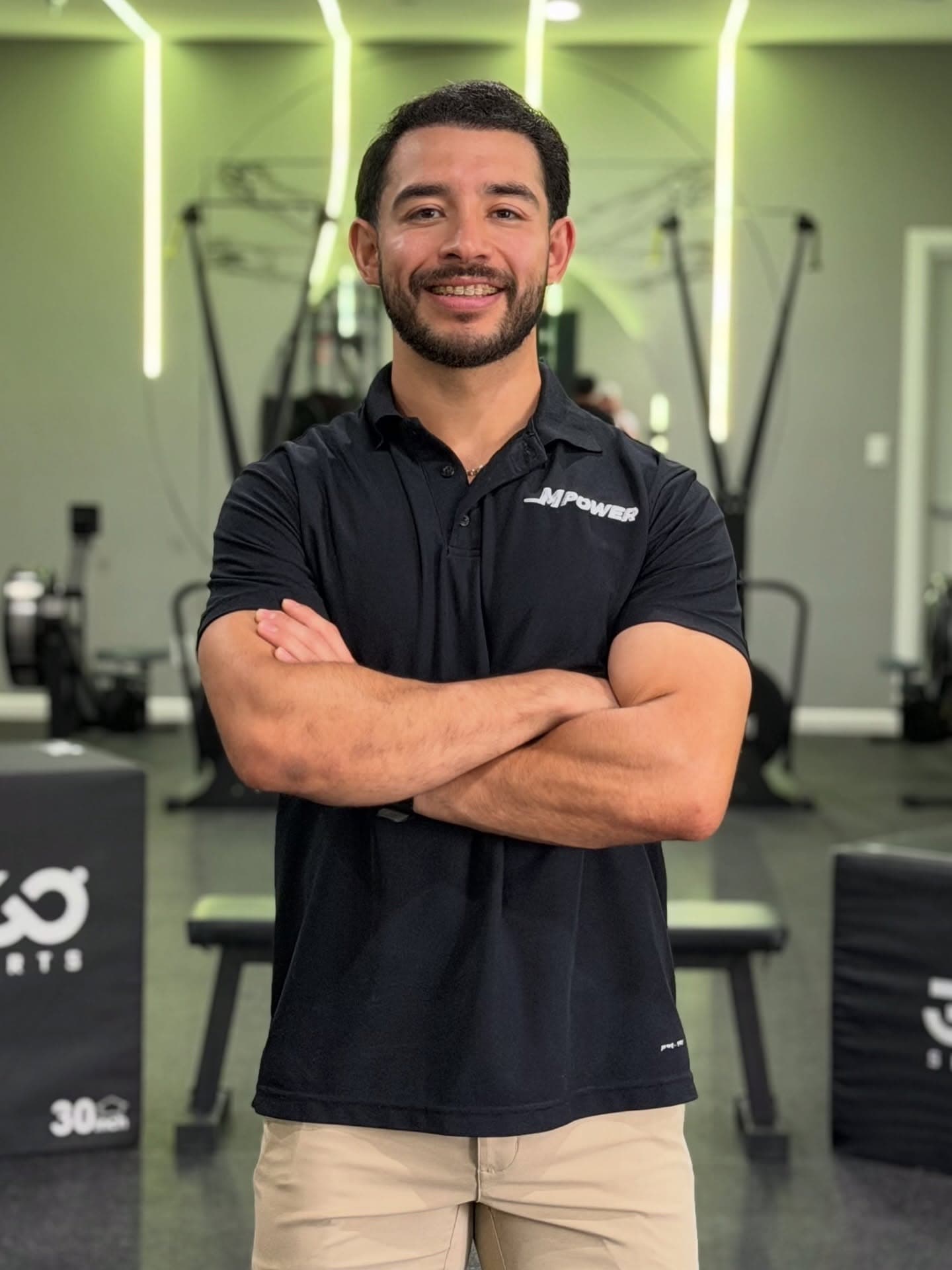 An Mpower coach in a black studio polo, arms crossed on the training floor